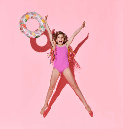 Happy girl with bright cornrows in one-piece rose swimsuit jumping and screaming full of joy, throwing rubber ring with fruit pattern. Full length shot isolated on pink. Childhood, emotions, summerの写真素材