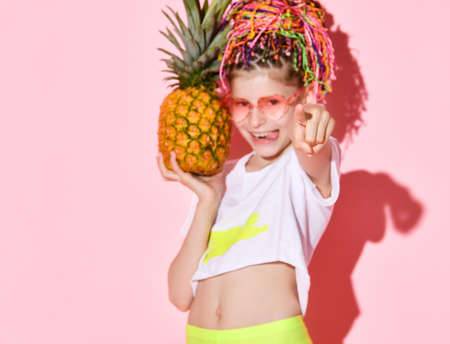 Active girl with rainbow colored African braids on her head holding pineapple in hand pointing at the camera with a cheerful smile. Cropped shot isolated on pink.の写真素材