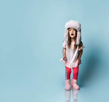 Little blonde kid with long hair, dressed in pink blouse, plush hat, pants and boots. She raised her...
