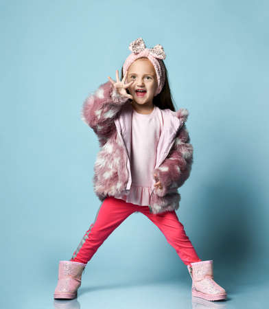 Little blonde kid with headband, dressed in pink faux fur coat, pants and boots. She smiling, gesticulating, posing on blue studio background. Childhood, fashion, advertising. Full length, copy spaceの写真素材