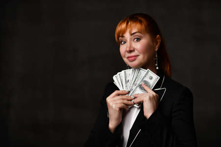 beautiful woman in a black jacket and white shirt looks at the camera with a charming smile, holding many dollars in her hands. Waist up shot on dark gray background, copy space. lady boss, noirの写真素材