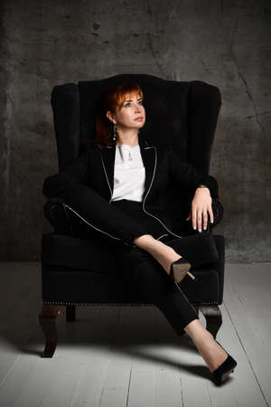 Thoughtful pretty woman in black trouser suit and high heels sits cross-legged in armchair at dark room pensively looking aside. Full length shot on grey background. Elegance, mystery, lady boss, noirの写真素材