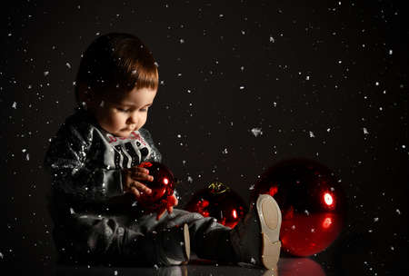 Baby girl in gray boots and sparkling suit. She is posing with three red balls, christmas decoration, sitting on floor in twilight, against black background. New Year, holidays. Snowing itの写真素材