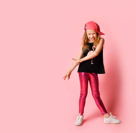 Funny little girl in stylish summer attire and baseball cap grimacing and lolling while looking at camera. Full length shot isolated on pink, copy space. Stylish, having fun, childhood, naughtyの写真素材