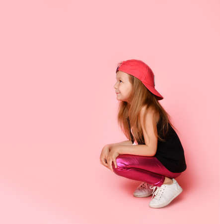 Charming active preschooler in trendy trendy dress, playful and smiling, squatting, looking to the side, on a pink background.の写真素材