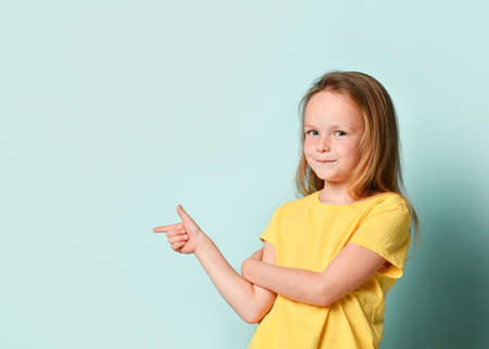 Cute blonde little girl in a yellow T-shirt. She smiles and points to the side with her index finger, looks at you. Presenting on a green studio background.の写真素材