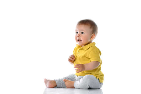 small baby in a yellow polo sits on the floor, laughs, touches his legs and looks to the side at the top corner on a white background. Happy childhood conceptの写真素材