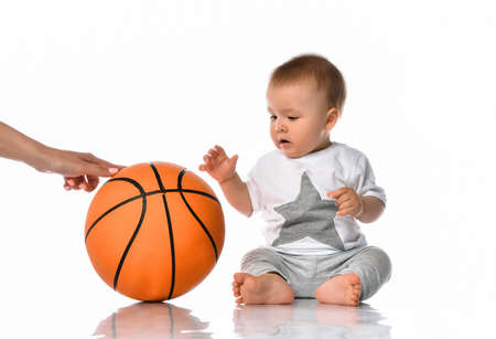 Cute baby boy in casual jumpsuit sitting on floor. Mother hand passing basketball ball to adorable toddler child. Sport game, activity and leisure. Studio portrait on white wall backgroundの写真素材
