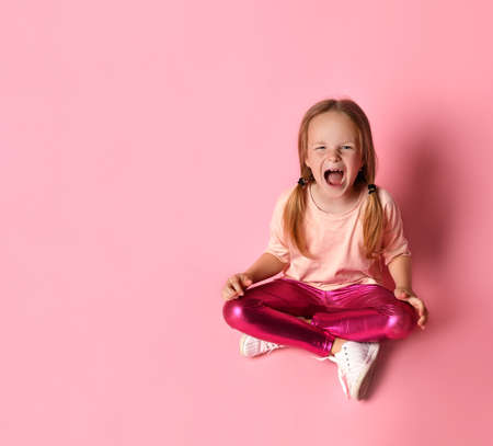 Little blond woman in a t-shirt, leggings and white sneakers. She sits on the floor and screams with her mouth wide open on a pink background. Childhood, emotions, gestures. Copy spaceの写真素材