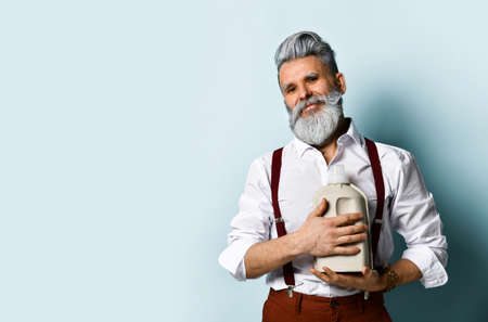 bearded man in white shirt, brown pants and suspenders, bracelet. Holds a plastic paper eco bottle, posing on a blue background. Close up, copy spaceの写真素材