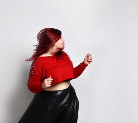 Fat red lady in a red top with spikes, a bra with a red top and a leather skirt, earrings. She is dancing and singing, posing on a white background photo. The concept of fashion and style. Close copy spaceの写真素材