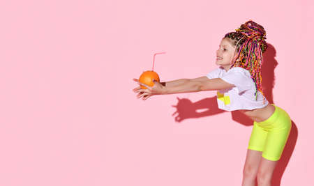 Cheerful schoolgirl in shorts and top leaning aside holding orange with swizzle stick in...