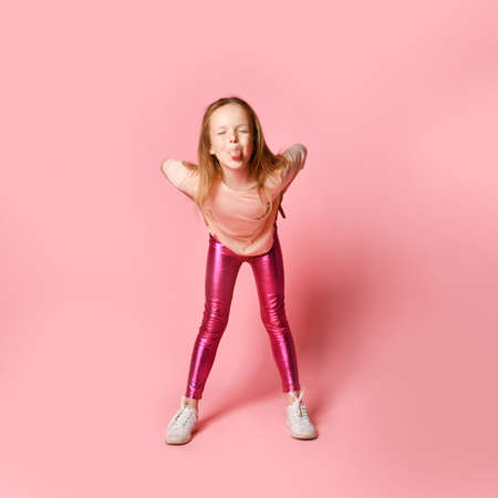Joyful preschool girl in stylish blouse and leggings is teasing someone leaning forward, grimacing and lolling. Full length shot isolated on pink. Stylish kids, happy childhood, emotions, games, funの写真素材