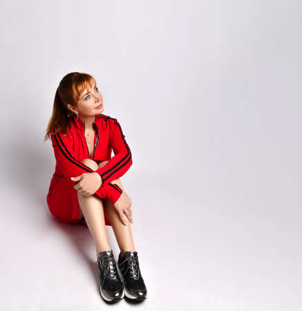 Pensive young girl in trendy outfit sitting on floor. Dreamy female wearing red suit fashionable sneakers posing in with hand around knees. Grey copy space. Top above high angle view full length photoの写真素材
