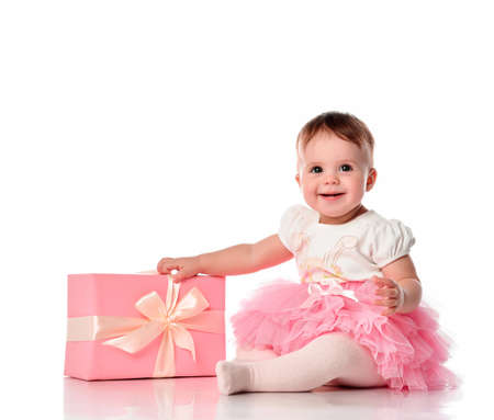 Happy smiling baby girl with wrapped gift box sitting on white studio floor isolated. Toddler child full length portrait. Congratulation, birthday party celebration. Positive emotionの写真素材