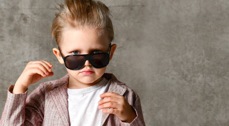 little boy fashionista in light jeans and a stylish jacket, looking over his glasses against the background of a concrete wall. Stylish childhoodの写真素材