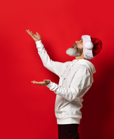 Side view of bearded man in santa hat catching snow standing on red background. Senior hipster stands under the free space for the text. Christmas and holiday discounts concept.の写真素材