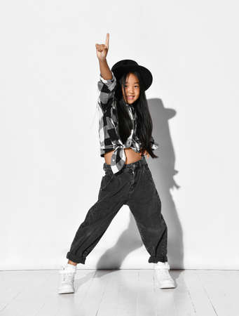 Portrait of an Asian girl with black hair who dances and has fun on a white background. The child wears a black hat, plaid shirt, jeans and looks into the camera. Children's fashion and style.の写真素材