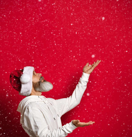 Winter emotion. Side view of bearded man in santa hat catching snow standing on red background. Senior hipster stands under the free space for the text. Christmas and holiday discounts concept.の写真素材