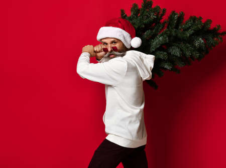 Glamorous senior man with a white beard and fashionable glasses, wearing a Santa hat, carries a Christmas tree on his shoulder for the holiday. on a burgundy background. christmas conceptの写真素材
