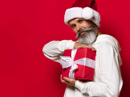 Senior bearded man holding a gift box on a red background and looking at the camera with a smile. Stylish and fashionable Santa Claus wishes you a Merry Christmas and a Happy New Year 2021.の写真素材