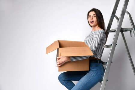 Surprised young woman holding a cardboard box on a light background, sitting on the iron stairs. We...
