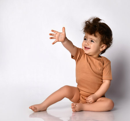 Cute little curly boy extends his hand forward sitting barefoot on a gray background.の写真素材