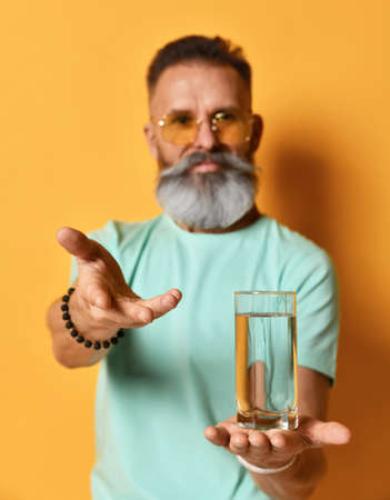 Close up of a glass of water in the hands of a bearded man standing against a orange wall.