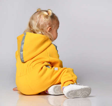 Little toddler girl in a yellow warm sports suit sitting on a gray background.の写真素材