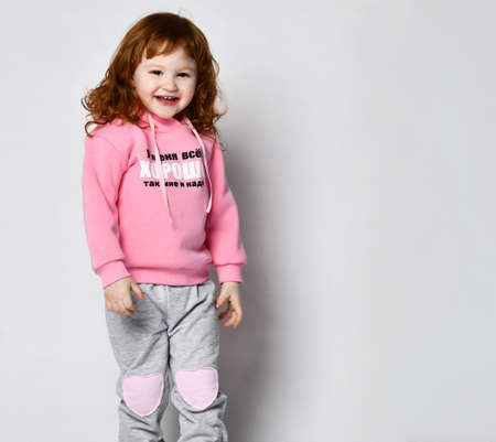 Isolated image of a cute adorable red-haired baby girl in a pink lettering hoodie posing in an empty...