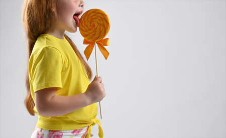 Cropped photo of a girl in a summer fashion outfit licking lollipops. Studio shot. Happy little female child biting sweet caramel candy on stick enjoying delicious taste.の写真素材