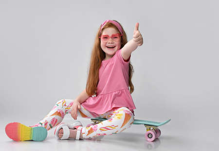 Smiling little red-haired girl child sitting on skateboard gesturing victory