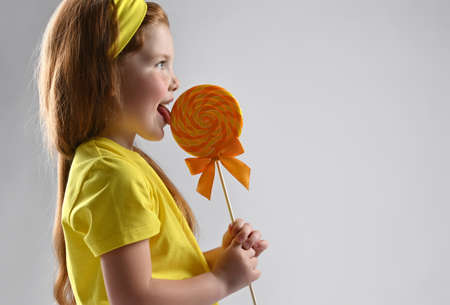 Smiling little red-haired girl licking lollipops side view studio shotの写真素材