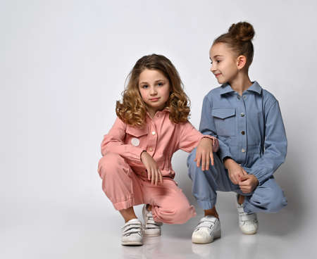 Two smiling girls, friends, sisters in blue and pink cotton overalls with pockets are squatting...