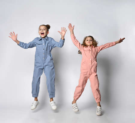 Two smiling girls, friends, sisters in blue and pink cotton overalls with pockets merrily jump and...
