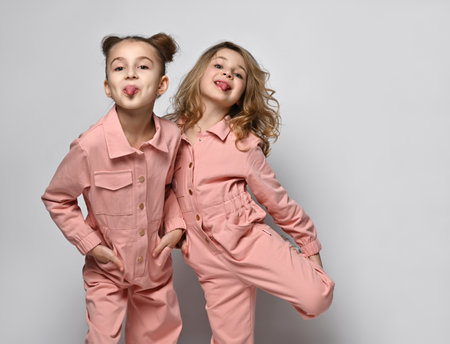 Two smiling girls, girlfriends, sisters in pink cotton overalls with pockets stand close to each other, put their hands in their pockets and provocatively show us their tonguesの写真素材
