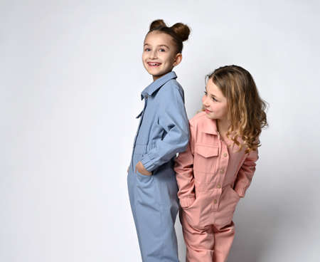 Two smiling girls, girlfriends, sisters in blue and pink cotton overalls with pockets stand close to each other with their backs, one looks at the camera, the other looks to the side.の写真素材