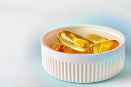Yellow softgels or capsules are in bottle cap. fish oil. White and blue surface. Vitamins and healthy lifestyle. Bright background about health and medicine with copy space. Softgel close-up. Macroの写真素材