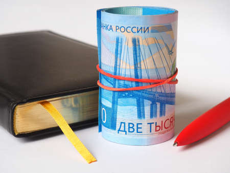 Black diary, red pen and bills of two thousand rubles. Banknotes rolled into a tube and tied with an elastic band. The banknotes depict the bridge to Russky island in Vladivostokの写真素材