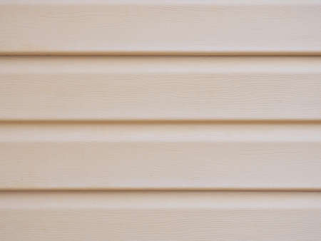A wall of a house sheathed with plastic siding close-up. Light beige brown background or wallpaper. The material is faded and not new. reduced contrast. Construction and repair of buildingsの写真素材