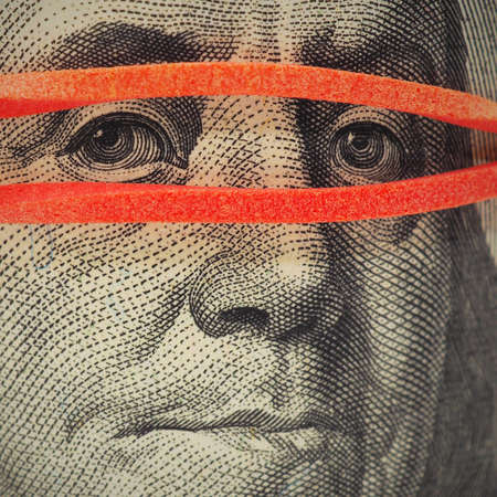 A 100 dollar banknote is very close up. Franklin's face is covered with a rubber band like a mask....