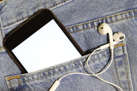 Template for audio and video content. Phone with a white screen and headphones in a jeans pocket. Bright photo to insert your image.の写真素材