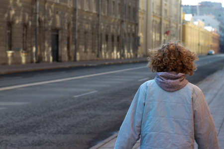 Girl with curly blond hair walks in city street near the road. Copy space outdoor. Photo from the back.の写真素材