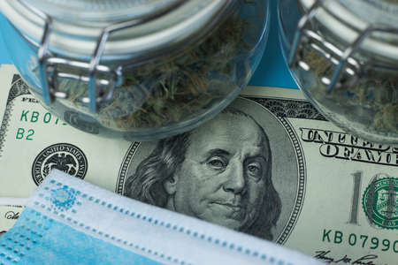 One hundred dollars banknote close-up. Cannabis in jars and respiratory safe mask.