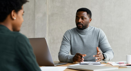 Man and a woman are sitting at a table with laptops in front of them. The man is talking to the woman, possibly discussing work or a project. The laptops are openの素材