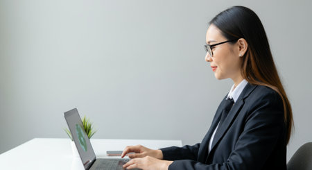 Woman is sitting at a desk and typing on a laptop. She is wearing a black suit and glassesの素材