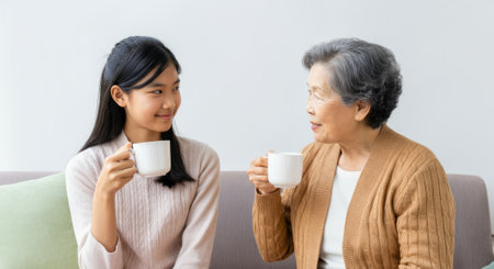 Woman in a white shirt is sitting next to an older woman in a brown sweater. Both women are holding cupsの素材