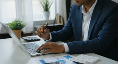 Man in a suit is typing on a laptop. He is using a pen to write on a piece of paperの素材