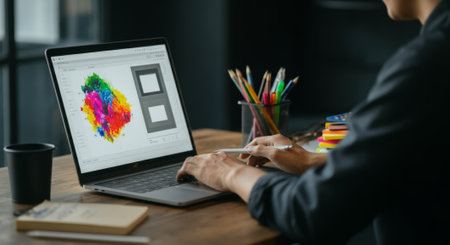 Person is using a laptop computer. The laptop is open and has a colorful image on the screen. There are several pens and pencils on the desk, and a cup is nearbyの素材