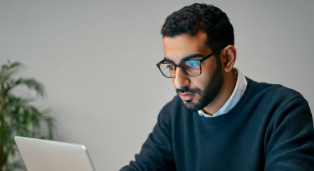 Man wearing glasses is sitting at a desk with a laptop. He is wearing a black sweater and a white shirtの素材